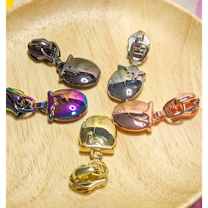 May include: Four zipper pulls in different colours and finishes. The pulls are in the shape of a small bucket with a loop on top. The colours are gold, silver, rose gold, and iridescent.