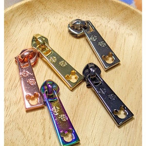 May include: Four zipper pulls in different finishes: gold, rose gold, black and iridescent. Each pull features a Mickey Mouse head cutout and a snowflake design.