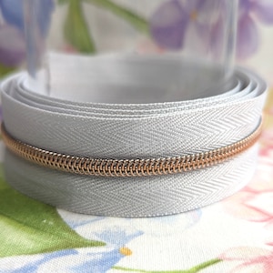 May include: A roll of pale grey zipper tape with a copper-coloured metal zipper. The tape has a herringbone weave pattern. The background is a soft floral print.