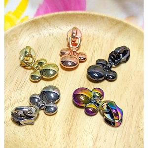 May include: Five zipper pulls in different colors, including gold, silver, black, rose gold, and iridescent. Each pull features a three-dimensional Mickey Mouse head design.