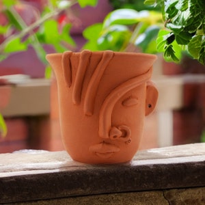 May include: A terracotta planter shaped like a face with a stylized hair design. The planter has a brown glaze and a small hole in the ear.