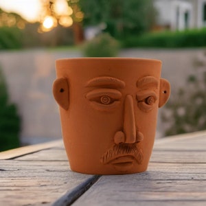 May include: A terracotta planter shaped like a face with a moustache and a serious expression. The planter has a brown glaze and is made of clay.
