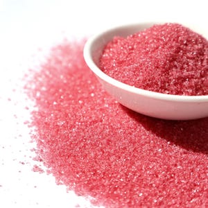 Könnte beinhalten: Eine weiße Keramikschale gefüllt mit leuchtend rosa kristallinem Zucker, wobei etwas Zucker auf eine weiße Oberfläche verschüttet wurde. Die Zuckerkristalle glitzern und erzeugen einen auffälligen Kontrast zum schlichten Hintergrund.