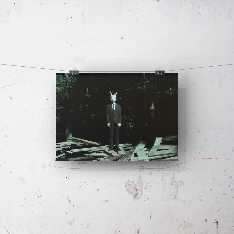 May include: A person wearing a white rabbit mask and a dark suit stands in a dark forest setting. The person is standing on a pile of wooden planks.