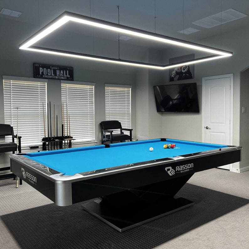 K&ouml;nnte beinhalten: Ein blauer Billardtisch mit schwarzen Verzierungen und einem schwarzen Untergestell. Der Tisch hat einen schwarzen Filzbezug und ein schwarz-wei&szlig;es Rasson Billiards Logo. Der Tisch steht in einem Raum mit einer wei&szlig;en Decke und grauen W&auml;nden. Im Hintergrund befinden sich zwei Fenster mit Jalousien und eine T&uuml;r. Der Raum hat auch ein Schild mit der Aufschrift "Pool Hall", das an der Wand h&auml;ngt.