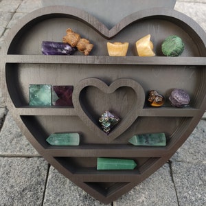 May include: A dark brown wooden heart-shaped display shelf with multiple shelves for displaying small objects. The shelves are filled with various colourful crystals and stones, including green, purple, and brown.