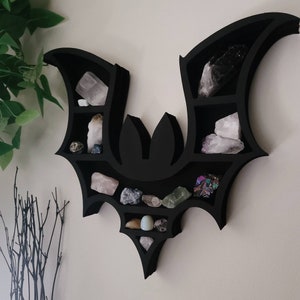 May include: Black bat-shaped wall shelf with multiple compartments for displaying crystals and other small objects. The shelf is mounted on a white wall.