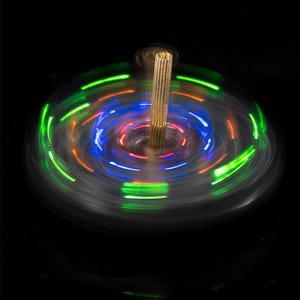 May include: A spinning top with a gold-colored central post and a swirling pattern of green, blue, and red lights. The toy is in motion, creating a blurred effect against a black background. The top is a classic toy.