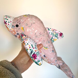 May include: A pink fabric dolphin toy with floral patterns and the text "Grow through what you go through." The dolphin has black button eyes and fins with colorful floral designs. The dolphin is being held by a person.