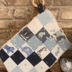 May include: A baby comfort blanket with a wooden ring and a patchwork design. The squares feature animal illustrations in blue and white, including a zebra, elephant, and giraffe. The blanket has a soft, white, fluffy border.