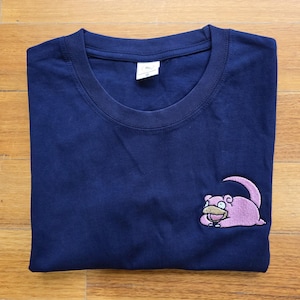 May include: A navy blue t-shirt featuring an embroidered pink Pokemon character on the front. The character is a pink, round creature with a long tail and a surprised expression.