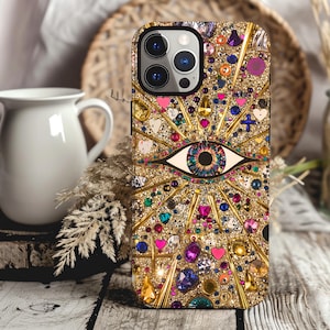 May include: A gold phone case with a large, colorful eye design. The eye is surrounded by rhinestones and other embellishments. The case is designed for an iPhone.