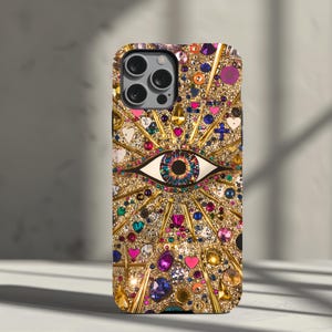 May include: A gold iPhone case with a colorful eye design and rhinestones. The eye is surrounded by a gold starburst pattern.
