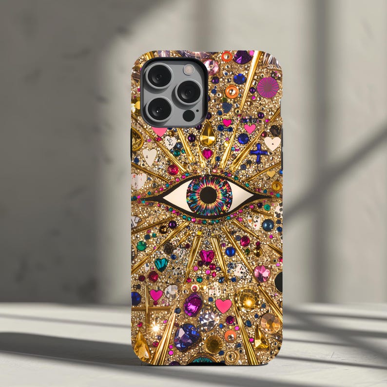 May include: A gold iPhone case with a colorful eye design and rhinestones. The eye is surrounded by a gold starburst pattern.
