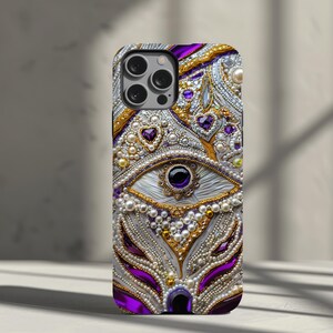 May include: A decorative phone case featuring an eye design with purple, gold, and white embellishments. The case is adorned with pearls, gems, and intricate detailing, creating a luxurious and ornate appearance. The case has a glossy finish.