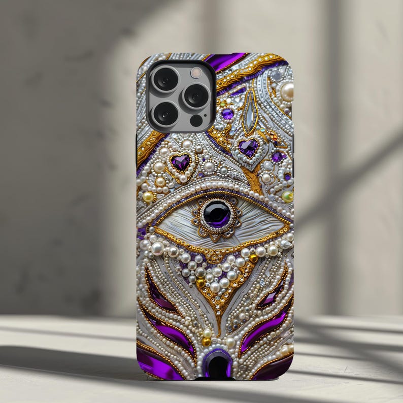 May include: A decorative phone case featuring an eye design with purple, gold, and white embellishments. The case is adorned with pearls, gems, and intricate detailing, creating a luxurious and ornate appearance. The case has a glossy finish.