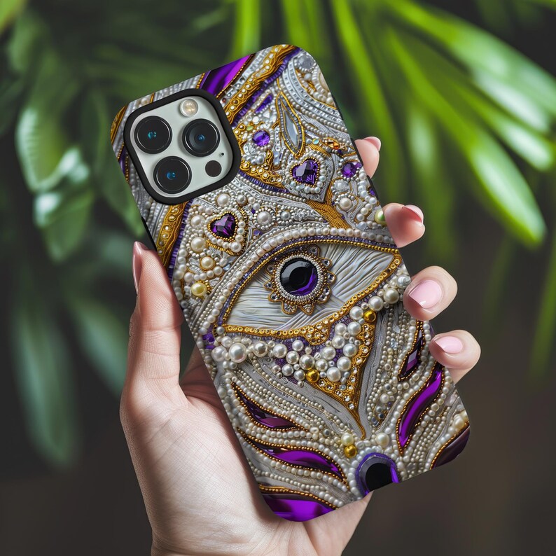 May include: A decorative phone case featuring an ornate design with white, gold, and purple embellishments. The case is adorned with pearls, heart-shaped gems, and a central eye motif. The phone case is held in a person's hand.
