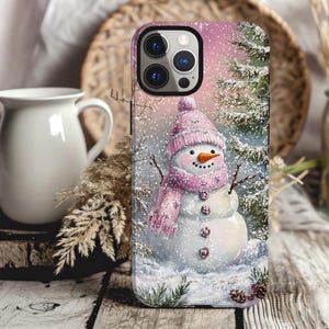 May include: A pink and white snowman with a pink scarf and a pink hat stands in a snowy winter scene. The background is a pink and white winter sky with a pine tree covered in snow. The image is on a phone case.