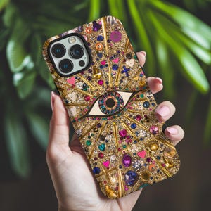 May include: A gold iPhone case with a large, colorful eye design. The eye is surrounded by rhinestones and glitter. The case has a black camera lens cutout.