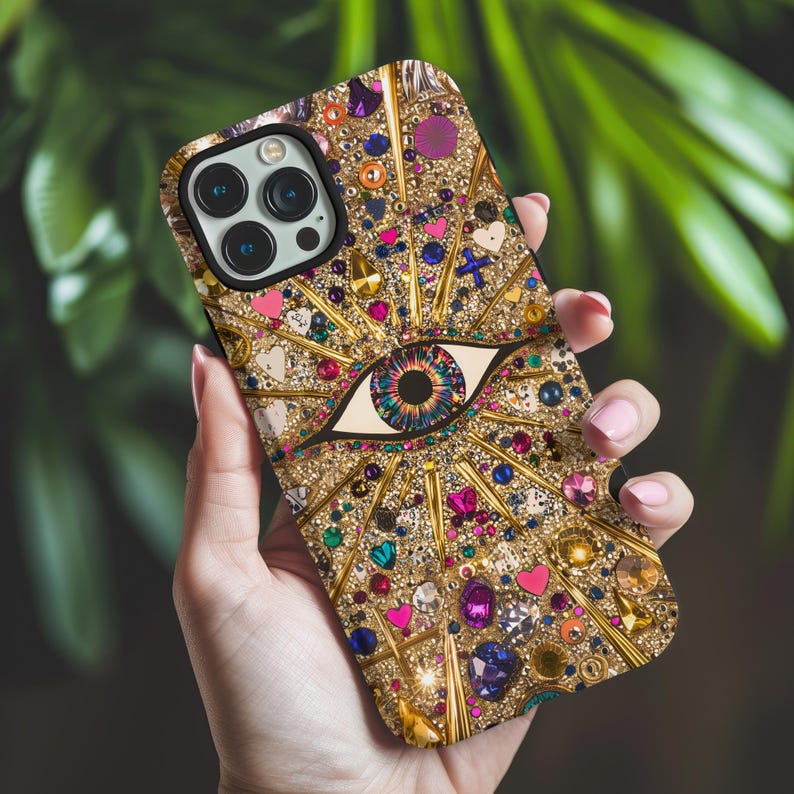 May include: A gold iPhone case with a large, colorful eye design. The eye is surrounded by rhinestones and glitter. The case has a black camera lens cutout.