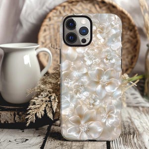 May include: A white iPhone case with a pearly, floral design. The case has a raised camera bump and a white border around the edges.