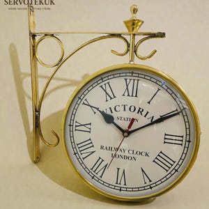 May include: A double-sided, gold-coloured railway clock with the text "VICTORIA STATION" and "RAILWAY CLOCK LONDON" on the white clock face. The clock features black Roman numerals and hands, and is mounted on a decorative gold bracket.