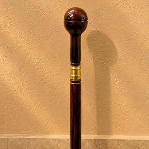 May include: A dark brown wooden walking stick featuring a polished spherical handle and a smooth, cylindrical shaft. A bright gold-colored metal band encircles the upper portion of the shaft, providing a distinct contrast to the deep wood grain.