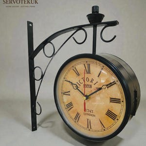 May include: A double-sided black metal clock with a vintage design. The clock face is tan with black Roman numerals and the words "VICTORIA STATION 1747 LONDON". The clock is mounted on a decorative black metal bracket, perfect for home decor.
