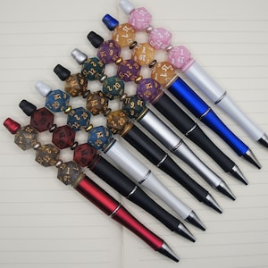 May include: A set of eight pens with different coloured dice attached to the barrel. The dice are numbered and have gold accents. The pens are all black with silver accents.