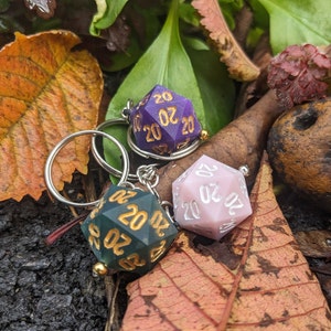 May include: Three 20-sided dice keychains, one purple, one pink, and one green, with gold numbers, on a bed of autumn leaves.