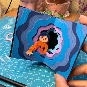 Puede incluir: Una obra de arte en papel hecha a mano con un diseño en capas y un recorte central. La obra representa un personaje con una camisa naranja y amarilla estampada, rodeado de capas onduladas azules y moradas, creando un efecto dimensional.