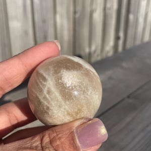 May include: A round, beige gemstone sphere with subtle white veining and a shimmering surface. The polished stone is held in a hand, showcasing its smooth texture and natural colour variations. The sphere is a decorative item.