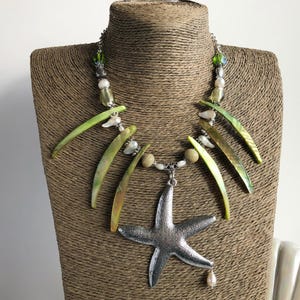 Könnte beinhalten: Eine grüne und weiße Muschelkette mit einem silbernen Seestern-Anhänger. Die Kette befindet sich auf einer braunen Mannequin-Büste.
