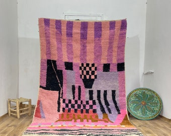 May include: A colorful Moroccan rug with a geometric pattern in shades of pink, purple, black, and white. The rug has a fringe border and is laid out on a wooden floor.