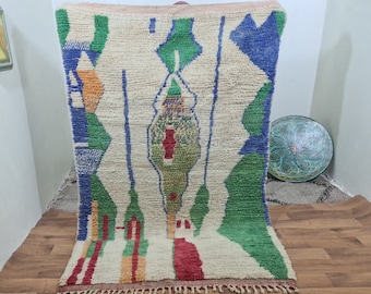 May include: A hand-woven Moroccan rug with a white background and colorful geometric patterns in shades of blue, green, red, and orange. The rug has a fringe border.
