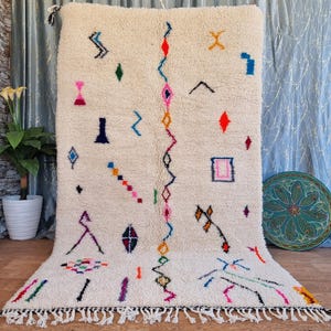 May include: A white Moroccan rug with a geometric pattern in various colors, including pink, blue, green, orange, and yellow. The rug has a fringe border and is made of wool.