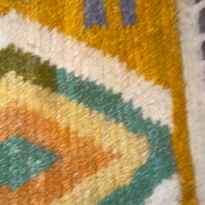 May include: A thick, textured rug featuring a geometric diamond pattern in shades of mustard yellow, sage green, and cream. The design includes two vertical blue rectangular accents near the top edge, with a shaggy pile surface throughout.