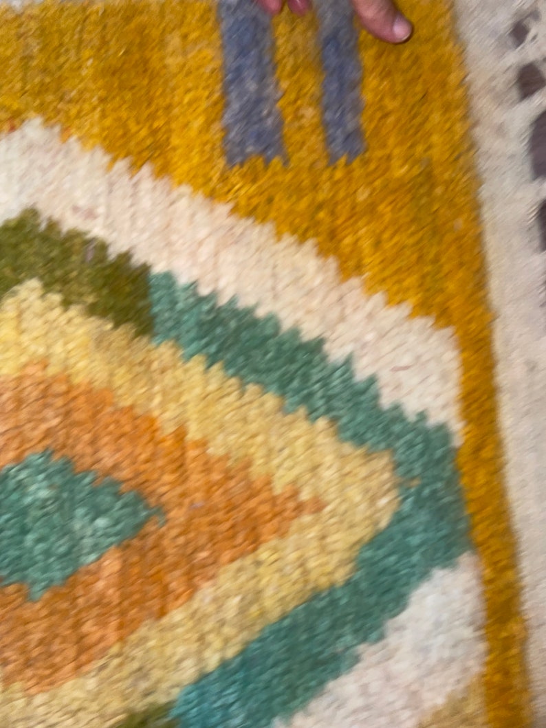May include: A thick, textured rug featuring a geometric diamond pattern in shades of mustard yellow, sage green, and cream. The design includes two vertical blue rectangular accents near the top edge, with a shaggy pile surface throughout.