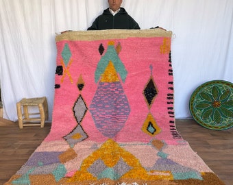 May include: A pink and multi-colored Moroccan rug with geometric patterns. The rug has a fringe border and is made of wool.