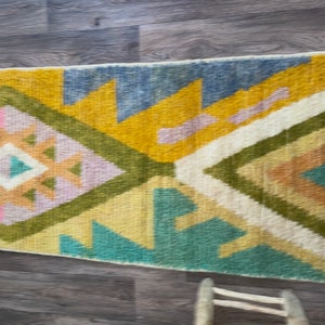 May include: A rectangular woven rug featuring a geometric pattern with large triangles and diamonds. The design incorporates a color palette of mustard yellow, sage green, slate blue, soft pink, and cream, arranged in a repeating symmetrical layout.