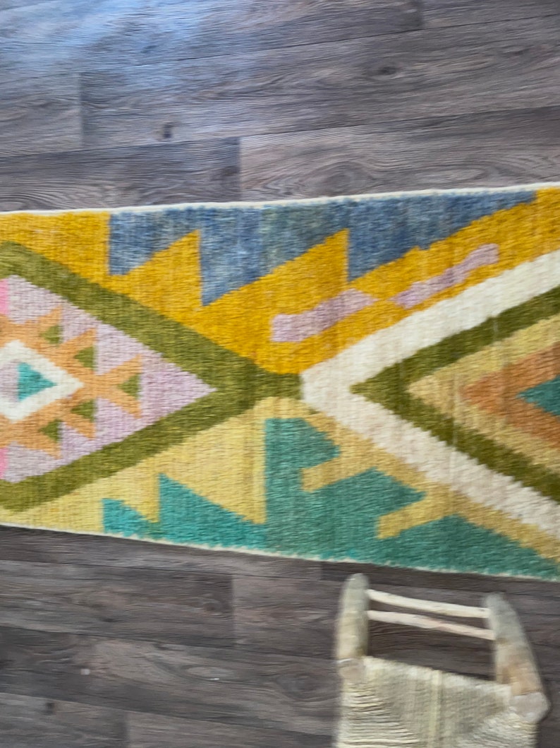 May include: A rectangular woven rug featuring a geometric pattern with large triangles and diamonds. The design incorporates a color palette of mustard yellow, sage green, slate blue, soft pink, and cream, arranged in a repeating symmetrical layout.