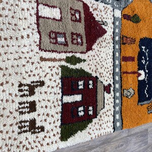 May include: A close-up of a colourful rug with a whimsical design featuring houses, trees, and a road. The rug is made of a soft, textured material and has a white background.
