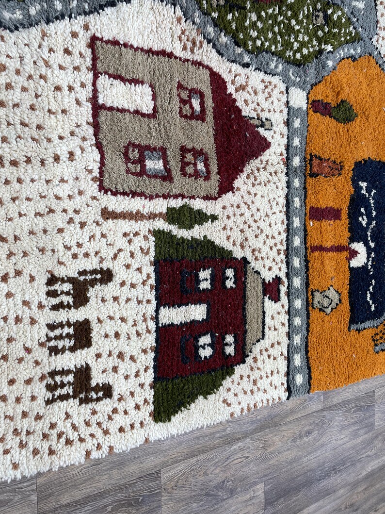 May include: A close-up of a colourful rug with a whimsical design featuring houses, trees, and a road. The rug is made of a soft, textured material and has a white background.