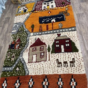 May include: A colourful Moroccan rug with a whimsical design featuring houses, trees, and animals. The rug has a cream background with orange, red, green, and brown accents. The border is a rich orange with black and white geometric patterns.