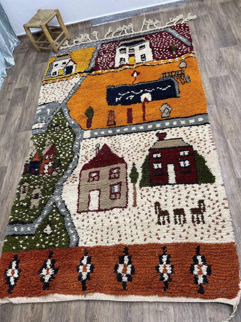 May include: A colourful Moroccan rug with a whimsical design featuring houses, trees, and animals. The rug has a cream background with orange, red, green, and brown accents. The border is a rich orange with black and white geometric patterns.