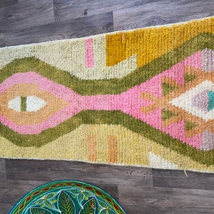 May include: A rectangular area rug featuring a high-pile texture with a geometric pattern in shades of mustard yellow, olive green, soft pink, and cream. Below it, a round, hand-painted green tray displays a central floral motif with leaves.