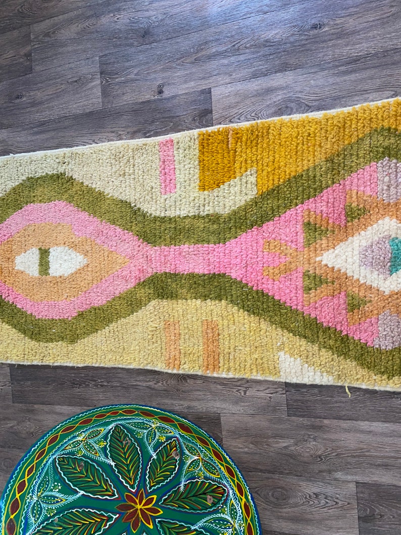 May include: A rectangular area rug featuring a high-pile texture with a geometric pattern in shades of mustard yellow, olive green, soft pink, and cream. Below it, a round, hand-painted green tray displays a central floral motif with leaves.
