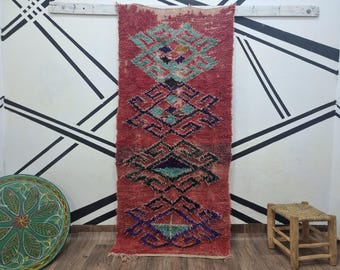 May include: A vintage Moroccan rug with a geometric pattern in shades of red, blue, and green. The rug has a distressed look and is made of wool.