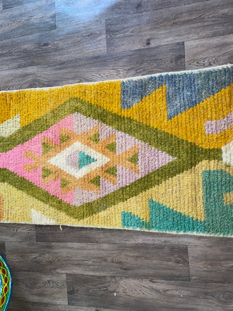 May include: A rectangular area rug featuring a high-pile texture with a central diamond motif. The design incorporates a vibrant color palette of mustard yellow, sage green, soft pink, light blue, and cream in a geometric pattern.