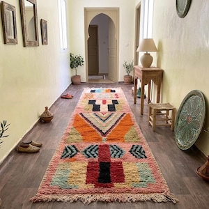 May include: A long, rectangular runner rug featuring a vibrant, multi-coloured geometric pattern with shades of pink, orange, black, green, and yellow. The rug has a thick, textured pile and is finished with fringed edges on both ends.